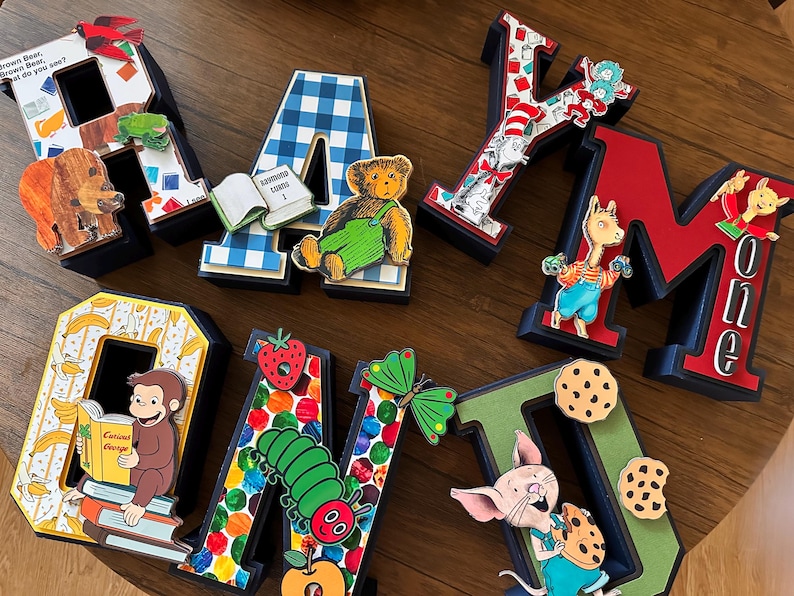 May include: A set of eight three-dimensional letters spelling RAYMOND, decorated with various children's book characters and motifs including a brown bear, a blue plaid pattern, a cat in a hat, a monkey, a caterpillar, and a pig eating cookies.