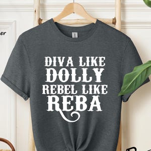 Puede incluir: Una camiseta de manga corta en color gris oscuro jaspeado con texto blanco impreso en el frente que dice DIVA LIKE DOLLY REBEL LIKE REBA. La prenda tiene cuello redondo, se muestra en una percha de madera y tiene el dobladillo anudado.