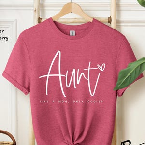 May include: A heather raspberry short-sleeve t-shirt featuring the white text "Aunt" in a script font with a small heart icon, followed by the text "LIKE A MOM, ONLY COOLER" in a simple sans-serif font centered on the front of the garment.