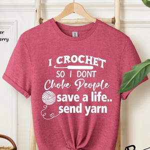 May include: A heather raspberry short-sleeve t-shirt with white text that reads I CROCHET SO I DONT Choke People save a life.. send yarn, featuring a graphic of a crochet hook and a ball of yarn with a loose thread ending in a small heart shape.