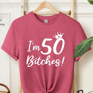 May include: A heather raspberry short-sleeve t-shirt with white screen-printed text that reads I'm 50 Bitches! with a small crown icon above the number 50. The shirt features a crew neckline and is displayed on a wooden hanger.