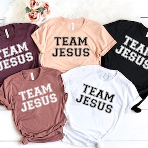 May include: Five t-shirts in different colors with the text "Team Jesus" printed in black with a white outline.