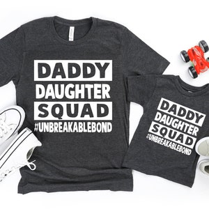 May include: Two gray t-shirts with white text. The larger shirt says "Daddy Daughter Squad #Unbreakablebond". The smaller shirt says "Daddy Daughter Squad #Unbreakablebond".