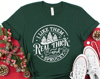 以下が含まれることがあります： 「I Like Them Real Thick and Sprucey」の白い文字が円形にデザインされた、濃い緑色のTシャツ。白いスニーカー、クリスマスオーナメント、お祝いの装飾が施されています。