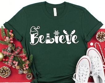 May include: A dark green t-shirt with the word "Believe" in white, featuring festive icons like a Santa hat, snowflake, and reindeer antlers. The shirt is styled with white sneakers, Christmas ornaments, and candy canes, creating a holiday theme.