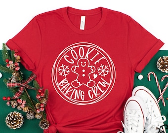 May include: A red t-shirt featuring a white circular design with the text "COOKIE BAKING CREW" and a gingerbread man. The image includes white trainers, candy canes, and festive decorations, suggesting a Christmas theme.