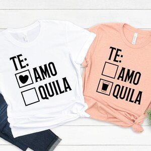 May include: Two white t-shirts with black text. The text on the left shirt says "TE: AMO QUILA" with a heart and a square checkbox. The text on the right shirt says "TE: AMO QUILA" with a shot glass and a square checkbox.