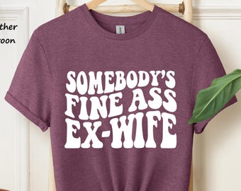 May include: A heather maroon short-sleeve t-shirt with white text printed on the front that reads SOMEBODY'S FINE ASS EX-WIFE in a bold, wavy, retro-inspired font. The shirt features a crew neckline and is displayed on a wooden hanger.