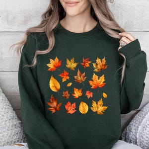 May include: A forest green crewneck sweatshirt featuring a central graphic print of various autumn leaves in shades of orange, yellow, red, and brown. The text Forest Green is printed in the upper left corner of the image.