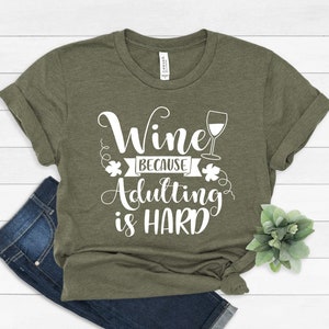 Pu&ograve; includere: T-shirt verde oliva con la scritta bianca "Wine because Adulting is HARD". Il design include un calice e decorazioni. La maglietta &egrave; piegata con un paio di jeans.