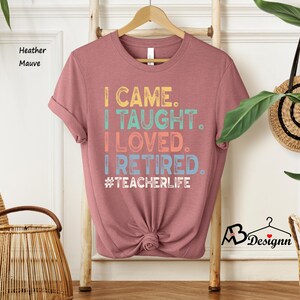 Może przedstawiać: Koszulka w kolorze wrzosowym z napisem "I CAME. I TAUGHT. I LOVED. I RETIRED. #TEACHERLIFE" w kolorowej, retro czcionce. Koszulka jest prezentowana na drewnianym wieszaku na neutralnym tle.