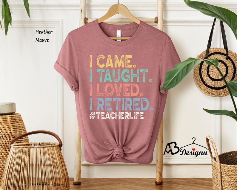 Może przedstawiać: Koszulka w kolorze wrzosowym z napisem "I CAME. I TAUGHT. I LOVED. I RETIRED. #TEACHERLIFE" w kolorowej, retro czcionce. Koszulka jest prezentowana na drewnianym wieszaku na neutralnym tle.