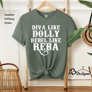 Puede incluir: Una camiseta de manga corta en color verde militar jaspeado con el texto en blanco DIVA LIKE DOLLY REBEL LIKE REBA. La prenda tiene cuello redondo, est&aacute; colgada en una percha de madera y tiene el dobladillo inferior atado en un nudo.