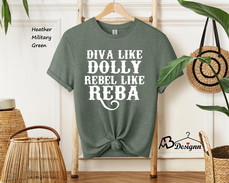 Puede incluir: Una camiseta de manga corta en color verde militar jaspeado con el texto en blanco DIVA LIKE DOLLY REBEL LIKE REBA. La prenda tiene cuello redondo, est&aacute; colgada en una percha de madera y tiene el dobladillo inferior atado en un nudo.