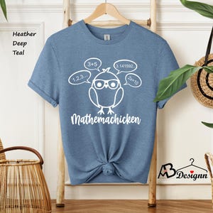 Op de afbeelding: Een blauw T-shirt met een witte afbeelding van een kip met een bril, omringd door wiskundige vergelijkingen. De tekst "Mathemachicken" staat onder de kip.