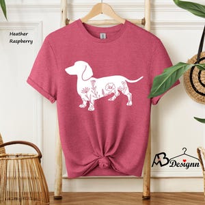May include: A heather raspberry t-shirt featuring a white outline of a dachshund with floral details. The shirt is folded and displayed on a wooden hanger.