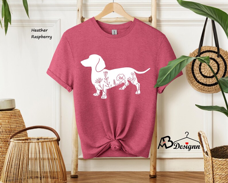 May include: A heather raspberry t-shirt featuring a white outline of a dachshund with floral details. The shirt is folded and displayed on a wooden hanger.