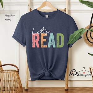 May include: A heather navy blue t-shirt with the text "Let's Read" in a colorful, distressed font. The text is in shades of pink, blue, green, and yellow.