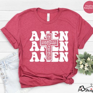 May include: A heather raspberry t-shirt with a white graphic of the word "Amen" repeated three times with a cross in the center. The cross is filled with a white floral pattern.