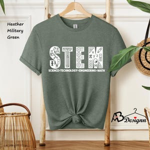 May include: A heather military green t-shirt with a white graphic of the word "STEM" made up of icons related to science, technology, engineering, and math. The text "SCIENCE TECHNOLOGY ENGINEERING MATH" is written below the graphic.
