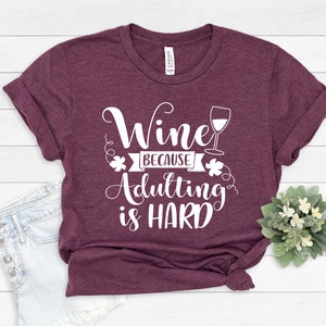 Pu&ograve; includere: Una maglietta bordeaux con le maniche arrotolate. La maglietta presenta il testo bianco "Wine because adulting is HARD" e un calice di vino. Un piccolo bouquet di fiori bianchi e verde &egrave; nell'angolo in basso a destra.
