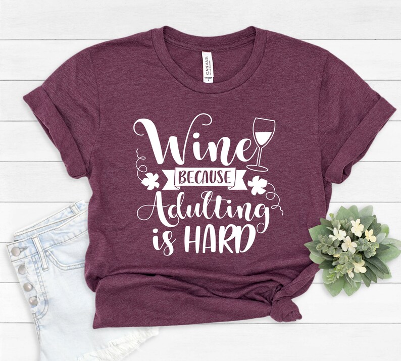 Pu&ograve; includere: Una maglietta bordeaux con le maniche arrotolate. La maglietta presenta il testo bianco "Wine because adulting is HARD" e un calice di vino. Un piccolo bouquet di fiori bianchi e verde &egrave; nell'angolo in basso a destra.