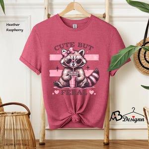 Op de afbeelding: Heather framboos t-shirt met een schattig wasbeer die een hartvormige zonnebril draagt en een roze drankje drinkt. Het shirt heeft de tekst "Cute But Fierce" met roze hartjes.