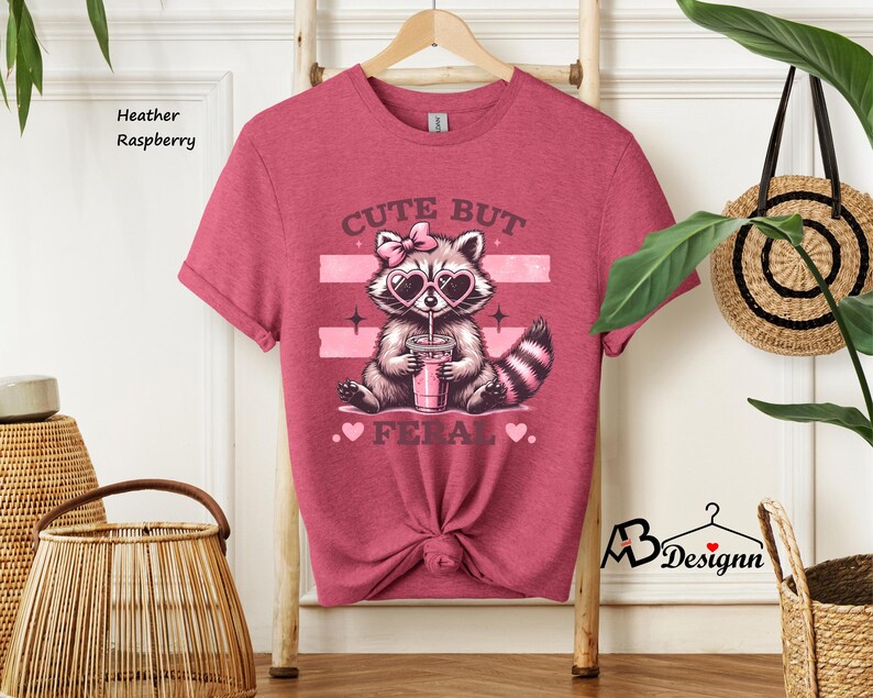 Op de afbeelding: Heather framboos t-shirt met een schattig wasbeer die een hartvormige zonnebril draagt en een roze drankje drinkt. Het shirt heeft de tekst "Cute But Fierce" met roze hartjes.