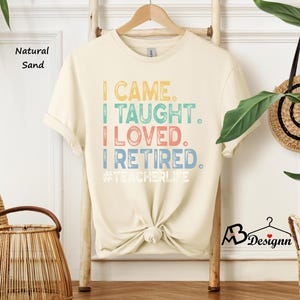 Może przedstawiać: Kremowa koszulka z napisem "I CAME. I TAUGHT. I LOVED. I RETIRED." w kolorowych literach. Koszulka zawiera r&oacute;wnież hashtag #TEACHERLIFE. Koszulka wisi na drewnianym wieszaku.