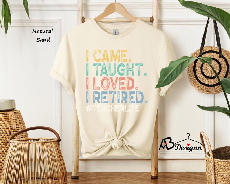 Może przedstawiać: Kremowa koszulka z napisem "I CAME. I TAUGHT. I LOVED. I RETIRED." w kolorowych literach. Koszulka zawiera r&oacute;wnież hashtag #TEACHERLIFE. Koszulka wisi na drewnianym wieszaku.