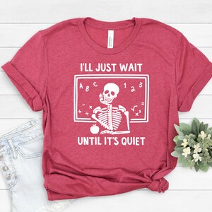 Może przedstawiać: Czerwony T-shirt z białym nadrukiem szkieletu. Szkielet siedzi przy tablicy z literami A, B, C i liczbami 1, 2, 3. Tekst na koszulce brzmi "Po prostu poczekam, aż będzie cicho".