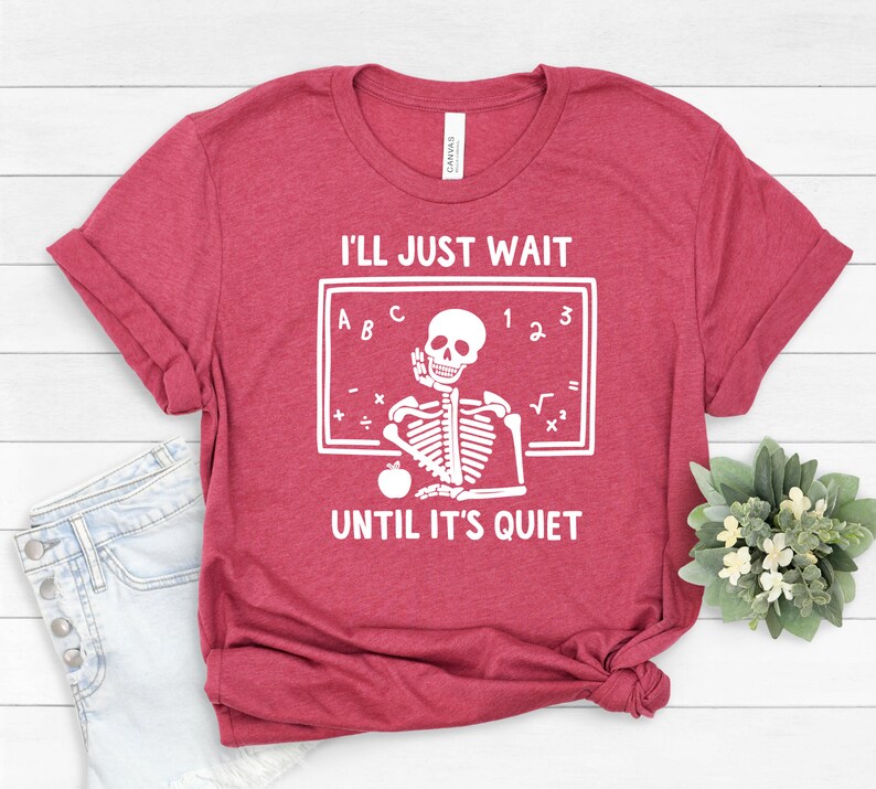 Może przedstawiać: Czerwony T-shirt z białym nadrukiem szkieletu. Szkielet siedzi przy tablicy z literami A, B, C i liczbami 1, 2, 3. Tekst na koszulce brzmi "Po prostu poczekam, aż będzie cicho".