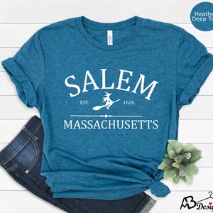 May include: A teal blue t-shirt with white text that reads "SALEM EST. 1626. MASSACHUSETTS" with a witch flying on a broomstick between the words "EST." and "1626."