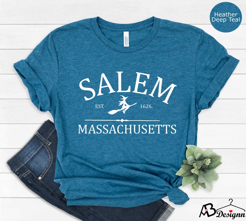 May include: A teal blue t-shirt with white text that reads "SALEM EST. 1626. MASSACHUSETTS" with a witch flying on a broomstick between the words "EST." and "1626."