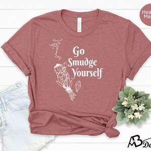 K&ouml;nnte beinhalten: Ein T-Shirt in der Farbe Heide-Mauve mit dem Text "Go Smudge Yourself" in Wei&szlig;. Das Shirt zeigt eine wei&szlig;e Illustration eines R&auml;ucherstabs mit aufsteigendem Rauch.