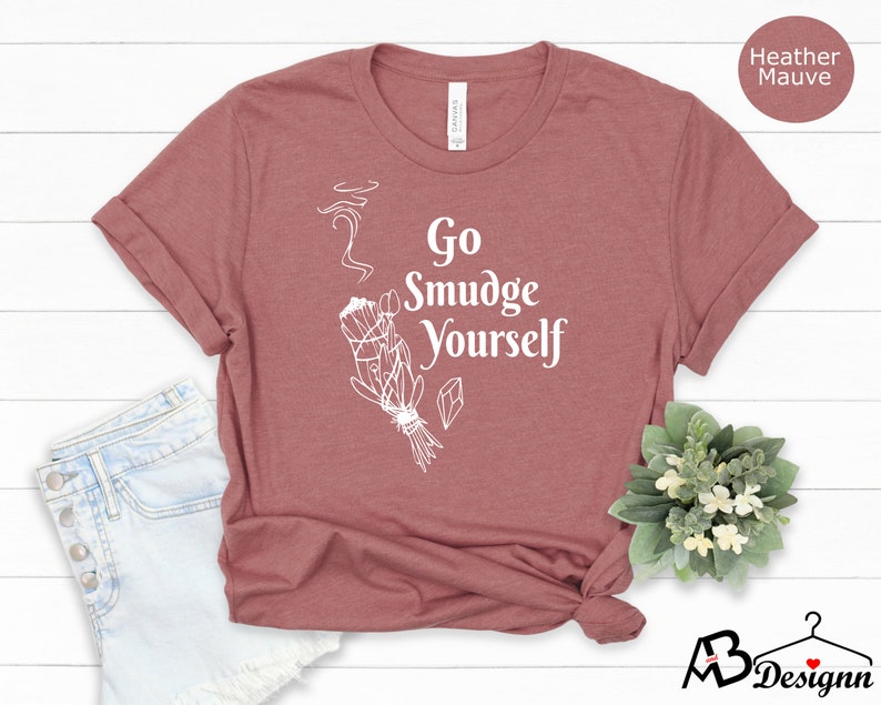 K&ouml;nnte beinhalten: Ein T-Shirt in der Farbe Heide-Mauve mit dem Text "Go Smudge Yourself" in Wei&szlig;. Das Shirt zeigt eine wei&szlig;e Illustration eines R&auml;ucherstabs mit aufsteigendem Rauch.