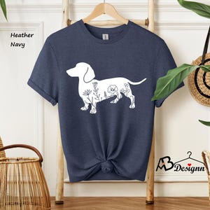 May include: A heather navy blue t-shirt featuring a white outline of a dachshund with floral details. The text "Heather Navy" is visible in the top left corner of the image.