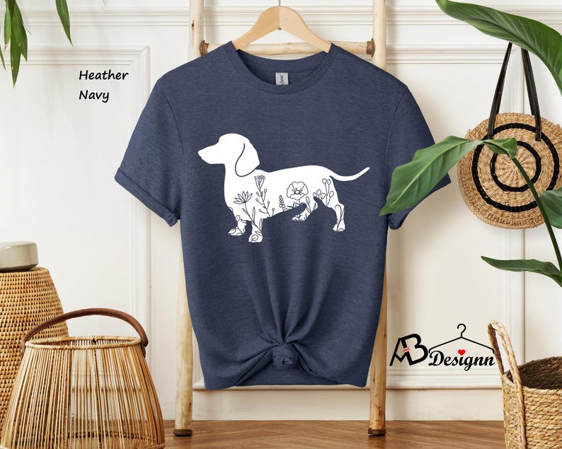 May include: A heather navy blue t-shirt featuring a white outline of a dachshund with floral details. The text "Heather Navy" is visible in the top left corner of the image.