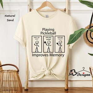 May include: A beige t-shirt with black text that reads "Playing Pickleball Improves Memory". The text is accompanied by three stick figure illustrations. The first stick figure is asking "What's the score?", the second is asking "Who just served?" and the third is asking "Were you 1 or 2?"