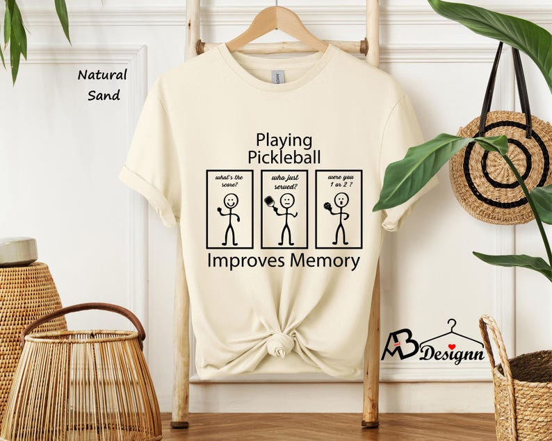 May include: A beige t-shirt with black text that reads "Playing Pickleball Improves Memory". The text is accompanied by three stick figure illustrations. The first stick figure is asking "What's the score?", the second is asking "Who just served?" and the third is asking "Were you 1 or 2?"