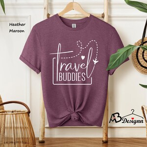 K&ouml;nnte beinhalten: Ein kurz&auml;rmeliges T-Shirt in heather maroon mit wei&szlig;em Grafiktext, der travel BUDDIES liest, sowie einer gestrichelten Fluglinie, einem kleinen Herz und einem Flugzeugsymbol. Das T-Shirt h&auml;ngt an einem Holzkleiderb&uuml;gel und hat einen Knoten.