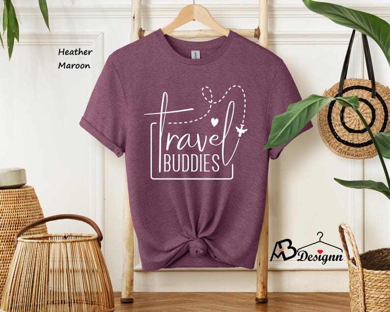 K&ouml;nnte beinhalten: Ein kurz&auml;rmeliges T-Shirt in heather maroon mit wei&szlig;em Grafiktext, der travel BUDDIES liest, sowie einer gestrichelten Fluglinie, einem kleinen Herz und einem Flugzeugsymbol. Das T-Shirt h&auml;ngt an einem Holzkleiderb&uuml;gel und hat einen Knoten.