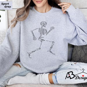 Peut inclure: Sweat-shirt gris sport avec un motif de coureur squelettique noir. Le motif utilise des termes anatomiques comme "radius" et "f&eacute;mur" pour former le squelette. Le sweat-shirt a un col rond et des manches longues.