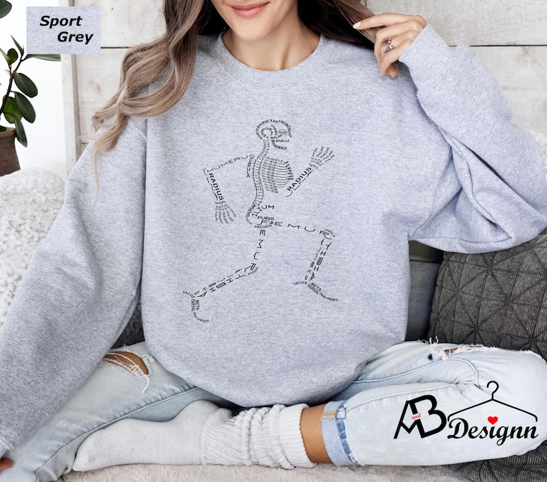 Peut inclure: Sweat-shirt gris sport avec un motif de coureur squelettique noir. Le motif utilise des termes anatomiques comme "radius" et "f&eacute;mur" pour former le squelette. Le sweat-shirt a un col rond et des manches longues.