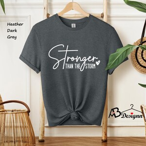 Może przedstawiać: Szary t-shirt w kolorze ciemnego melanżu z napisem "Stronger than the storm" w białym kolorze z małym symbolem serca.
