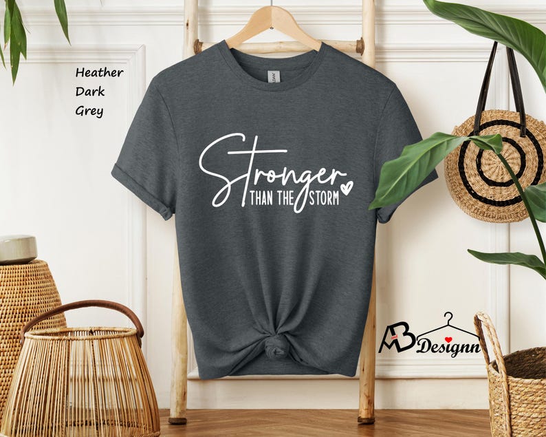 Może przedstawiać: Szary t-shirt w kolorze ciemnego melanżu z napisem "Stronger than the storm" w białym kolorze z małym symbolem serca.