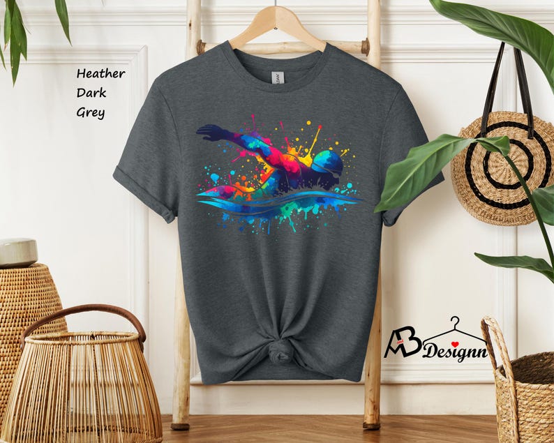 May include: A dark heather grey t-shirt with a colorful watercolor design of a swimmer. The design features splashes of blue, green, yellow, orange, and red.