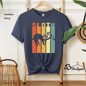 May include: Heather navy t-shirt with a retro design featuring the word "SLOTH" in orange and green block letters. A sloth illustration hangs from a branch. The shirt is knotted at the waist.