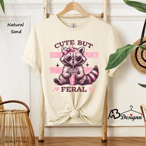 Op de afbeelding: Een cr&egrave;mekleurig T-shirt met een schattige wasbeer die een roze zonnebril en een roze strik draagt ​​en uit een roze beker drinkt. De tekst op het shirt luidt "Cute But Feral".