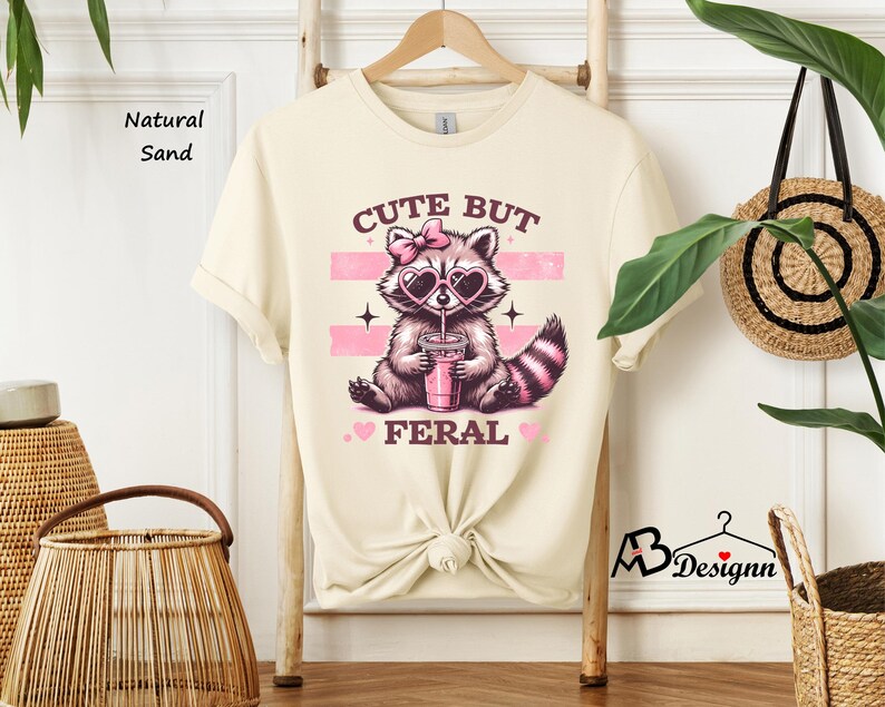 Op de afbeelding: Een cr&egrave;mekleurig T-shirt met een schattige wasbeer die een roze zonnebril en een roze strik draagt ​​en uit een roze beker drinkt. De tekst op het shirt luidt "Cute But Feral".